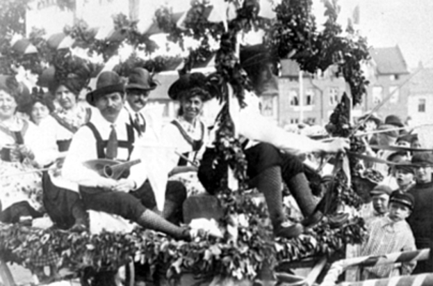Eine Gruppe von Menschen in traditioneller bayrischer Tracht fährt auf einemblattgeschmückten Festwagen bei einem Umzug mit, einige spielen Musikinstrumente, mit Gebäuden und einem klaren Himmel im Hintergrund, dargestellt in Schwarz-Weiß.