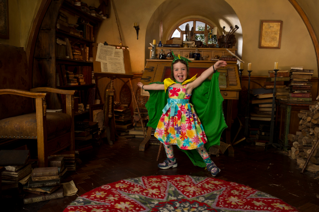 Ein Mädchen in einem Superman-Kostüm lächelt neben einem Tisch mit Büchern, Kerzen, Flaschen und anderen Gegenständen, mit einem Stuhl, Regalen, einem Fenster und einem Wandrahmen im Hintergrund.