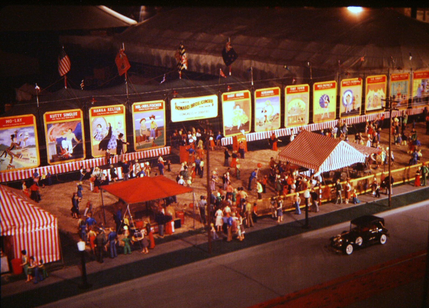 Altes Schwarz-Weiß-Foto eines nächtlichen Volksfestes mit Menschen, einem Auto, Masten mit Fahnen, einem Zaun, einem Zelt, Brettern mit Bildern und Text sowie einer beleuchteten Straßenlaterne.