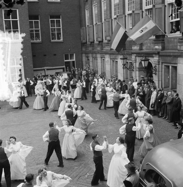 Eine Gruppe von Menschen, die auf der Straße vor einem Gebäude tanzen, mit einem geparkten Auto auf der rechten Seite und Fahnen und Lichtern, die an der Wand des Gebäudes befestigt sind, im Hintergrund abgebildet in Schwarz-Weiß.