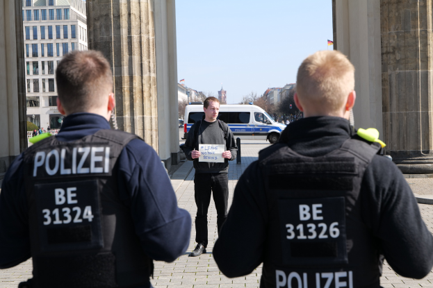 Drei Polizeibeamte stehen vor einem Gebäude, wobei ein Mann in der Mitte ein Papier hält und von Säulen flankiert wird, umgeben von Fahrzeugen, Menschen, Bäumen und Gebäuden unter dem Himmel.