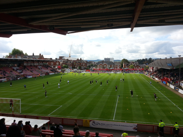 Ein Fußballspiel wird im St. Mary's Stadium gespielt, mit Zuschauern auf den Bänken und auf dem Boden stehend, einem Torpfosten auf der linken Seite und im Hintergrund Gebäuden, Bäumen und einem bewölkten Himmel.