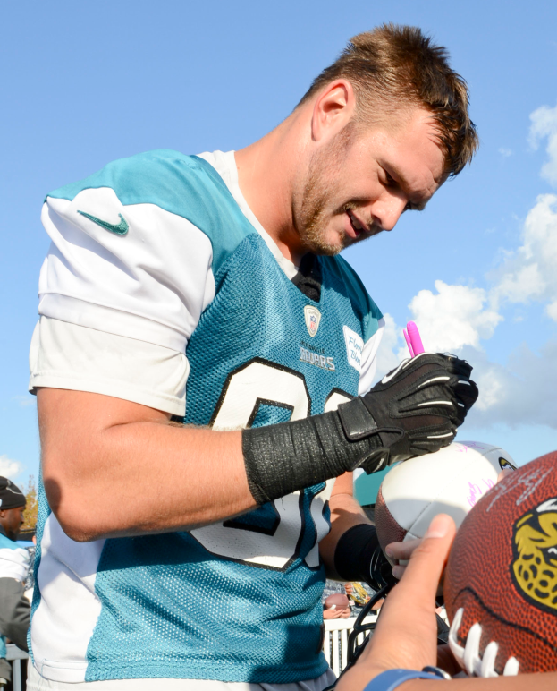 Jacksonville Jaguars Quarterback Blake Bortles in einem blauen und weißen Trikot signiert Autogramme für Fans, umgeben von einer Menge mit einigen, die Mützen tragen, unter einem Himmel voller weißer, flauschiger Wolken.