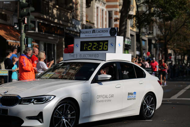Ein weißer BMW mit einer Uhr auf dem Dach fährt eine Straße entlang, mit einer Person im Auto und einer Gruppe von Menschen auf der Straße, während Gebäude, Bäume und Ampeln im Hintergrund zu sehen sind.