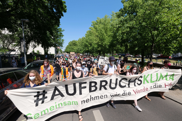 Eine Gruppe von Menschen marschiert auf einer von Bäumen gesäumten Straße, die eine Fahne mit der Aufschrift "Aufbrucksklima" schwenken, mit sichtbaren Gebäuden und geparkten Fahrzeugen.