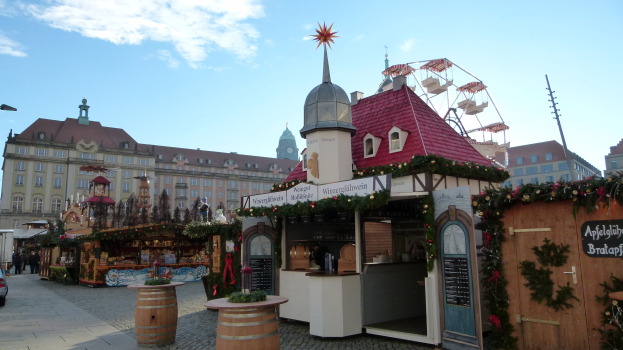 Ein geschäftiger Weihnachtsmarkt auf einem Stadtplatz mit geschmückten Bäumen, Gebäuden, Straßenlaternen, Fahrzeugen und Passanten unter einem bewölkten Himmel.