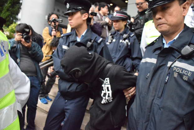 Eine Gruppe von Polizisten steht vor einer Menschenmenge, einige tragen Mützen und halten Kameras, mit einem Gebäude mit Säulen und Lichtern im Hintergrund und Bäumen auf der linken Seite.