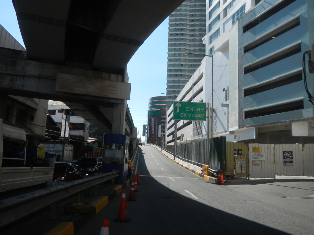 Stadtstraße mit Verkehrskegeln, Fahrzeugen, einer Brücke auf der linken Seite, Gebäuden mit Namensschildern, Laternenmasten und einem Himmel im Hintergrund.