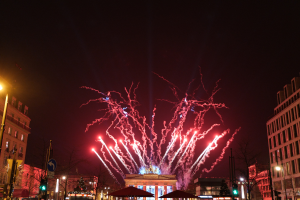 Eine belebte Stadtstraße in Berlin am Silvesterabend, voller Menschen, Fahrzeuge und festlicher Dekorationen, mit Feuerwerk, das den Himmel über den Gebäuden erhellt.