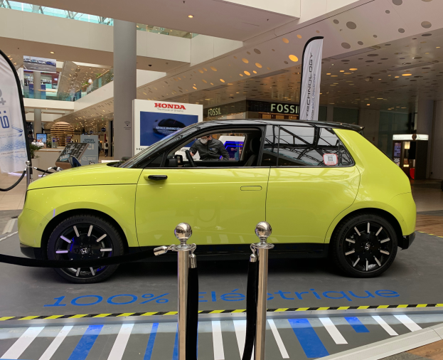 Ein gelbes wasserstoffbetriebenes Elektroauto steht drinnen auf einem Parkplatz, umgeben von Absperrpollern, Bannern und Menschen, mit Schildern, einem Topf mit Pflanze und Deckenleuchten im Hintergrund.