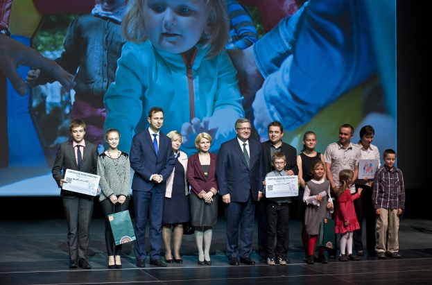 Eine Gruppe von Menschen auf einer Bühne mit Taschen und Urkunden, im Hintergrund ein Bildschirm mit dem Bild eines Kindes und anderen.