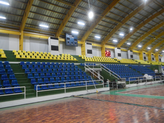 Eine geräumige Indoor-Sporthalle mit einem Holzboden, blauen und gelben Stühlen, Geländern, Treppen, die zu den Sitzplätzen führen, Bildschirmen an den Wänden, Deckenleuchten und einigen anwesenden Personen.