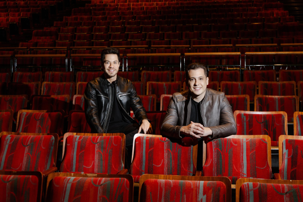 Zwei Männer in schwarzen Jacken, die während einer Vorstellung von *Dancing with the Stars* lächeln, während sie auf roten Auditoriumsstühlen sitzen.