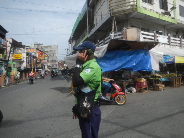 Ein Polizeibeamter in grüner und blauer Uniform und blauer Mütze steht an der Straße und telefoniert mit dem Handy, mit Fahrzeugen im Hintergrund, Ständen mit verschiedenen Gegenständen zu seiner Rechten und einem bewölkten Himmel.