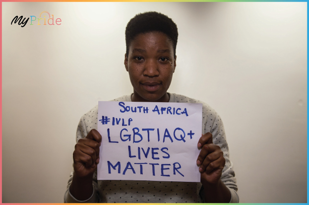 Eine Frau in einem weißen Hemd steht entschlossen vor einer Wand und hält ein Schild mit der Aufschrift "LGBTQ+ Lives Matter" in fetten schwarzen Buchstaben.