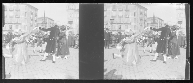 Ein Schwarz-Weiß-Foto einer Gruppe von Menschen, die auf der Straße während eines Faschingsumzugs tanzen, in Kostümen, mit Gebäuden, Straßenlaternen und -pfählen im Hintergrund bei klarem Himmel.