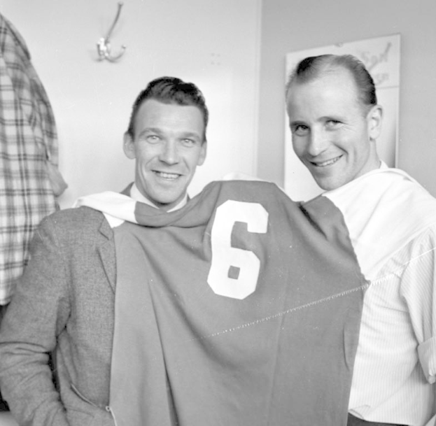 Zwei Männer in Schwarz und Weiß, die strahlend eine Fußballtrikot hochhalten, mit einer Tafel und einem Bügel an der Wand dahinter.