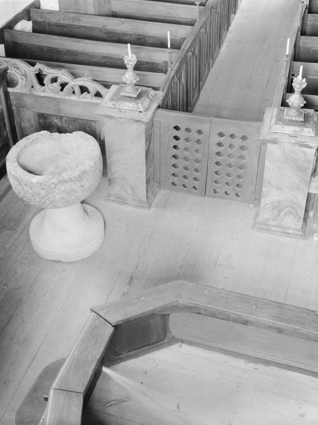 Ein Schwarz-Weiß-Foto einer Kircheninnenseite mit hölzernen Kirchenbänken, die in Reihen angeordnet sind, einer steinernen Schale in der Mitte und Kerzen auf Ständern auf beiden Seiten der Schale.