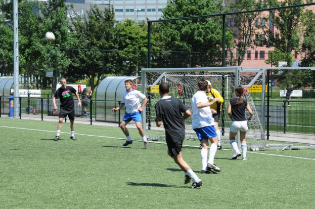 Personen in Kleidung beim Fußballspielen auf einem Feld mit einem Zaun und einem Tor in der Mitte, einem Pfosten und einem Fußball oben links und Bäumen und Gebäuden im Hintergrund.