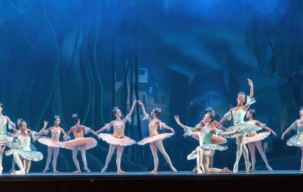 Eine Gruppe von Balletttänzern in bunten Kostümen, die *Der Nussknacker* auf der Bühne des Royal Opera House tanzen, sich synchronisiert gegen einen blauen Hintergrund bewegen.