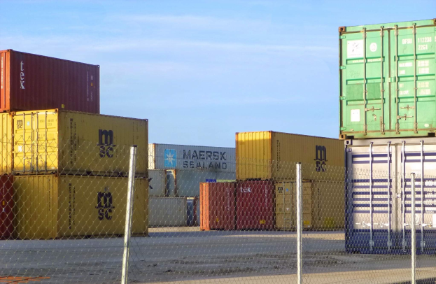 Ein dichter Stapel von Containern hinter Maschendraht, mit einer Straße unten und bewölktem Himmel im Hintergrund.