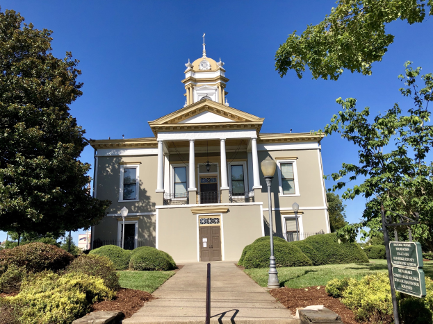 Großes Backsteingebäude mit einem Turm, umgeben von Fenstern, Türen, Säulen und Geländern, mit einem Schild, Straßenlaternen, Bäumen und einem bewölkten Himmel; das alte Rathaus hat kürzlich wiedereröffnet.