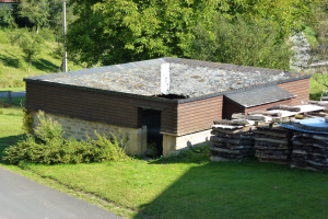 Kleines Holzhäuschen neben einer Straße mit Gras, Pflanzen, Bäumen, einem Pfahl und gestapelten Holzscheiten auf der rechten Seite.