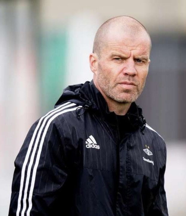 Bald male soccer coach in a navy blue jacket with white stripes and white collar standing on a field with clasped hands and a serious expression.