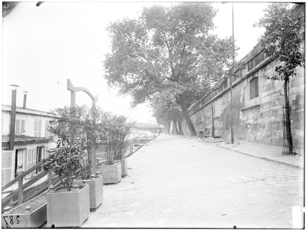 Ein Schwarz-Weiß-Foto einer Straße mit einer Wand auf der rechten Seite, Bäumen und Pflanzen auf der linken Seite und einer Brücke im Hintergrund, mit Text unten, der "New York City, New York, New Jersey, NY, USA, USA - Straßenansicht des New Yorker Staatsgefängnisses" lautet.