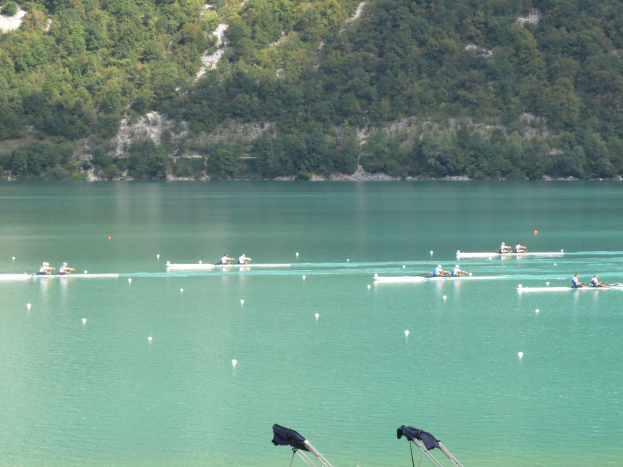 Eine Gruppe von Menschen, die bei einem Wettbewerb auf einem Gewässer mit Bäumen und einem Hügel im Hintergrund rudern, mit Kleidung, die über Tribünen am Boden drapiert ist.
