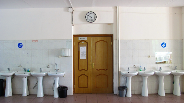 Reihe von Waschbecken mit Wasserhähnen, Mülltonnen auf dem Boden, eine Tür im Hintergrund, eine Uhr an der Wand und ein Spiegel auf der rechten Seite.