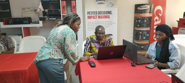 Eine Gruppe von Menschen sitzt um einen Tisch mit Laptops und Mobiltelefonen auf einer internationalen Geschäftsmesse in Senegal, mit Bannern, Schränken und einer Uhr im Hintergrund.