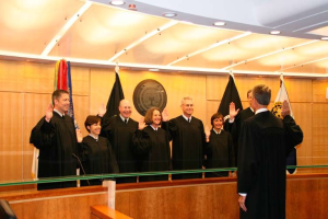 Eine Gruppe von Menschen in schwarzen Roben vor einem Gerichtssaal mit Flaggen an einer Holzwand, die unter Deckenlampen lächeln, mit einem Stuhl links und einem Richter in der Mitte.