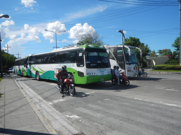 Ein grüner und weißer Shuttlebus steht am Straßenrand mit Motorrädern davor, ein grasbewachsener Fußweg links daneben und im Hintergrund Gebäude, Bäume und Laternenmasten unter einem klaren blauen Himmel.