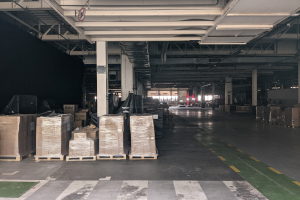 Großes Lagerhaus mit zahlreichen Kartonagenkisten auf Paletten, gestützt von Säulen und beleuchtet von Deckenlampen, mit einem an der Decke angebrachten Schild, das ein Flottenmanagementsystem anzeigt.