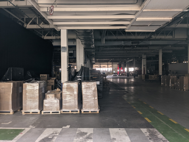 Großes Lagerhaus mit zahlreichen Kartonagenkisten auf Paletten, gestützt von Säulen und beleuchtet von Deckenlampen, mit einem an der Decke angebrachten Schild, das ein Flottenmanagementsystem anzeigt.