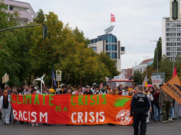 Eine Gruppe von Menschen marschiert auf einer von Bäumen gesäumten Straße, die eine Fahne mit der Aufschrift 'Klimakrise ist eine Krise' trägt, mit Gebäuden und einem klaren blauen Himmel im Hintergrund.