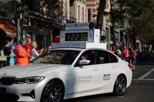 Ein weißer BMW mit einer Uhr auf dem Dach fährt eine Straße entlang, mit einer Person im Auto und einer Gruppe von Menschen auf der Straße, während Gebäude, Bäume und Ampeln im Hintergrund zu sehen sind.