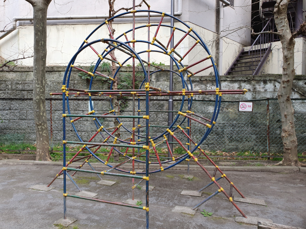 Ein Spielplatz mit einem großen Metallgerüst in der Mitte, umgeben von Bäumen und einer Wand mit Maschendrahtzaun, mit Treppen mit Geländern, die zu einem Gebäude mit Fenstern und Rohren führen, und einem Schild an einem Pfahl, alles auf einem grasbewachsenen Grund.
