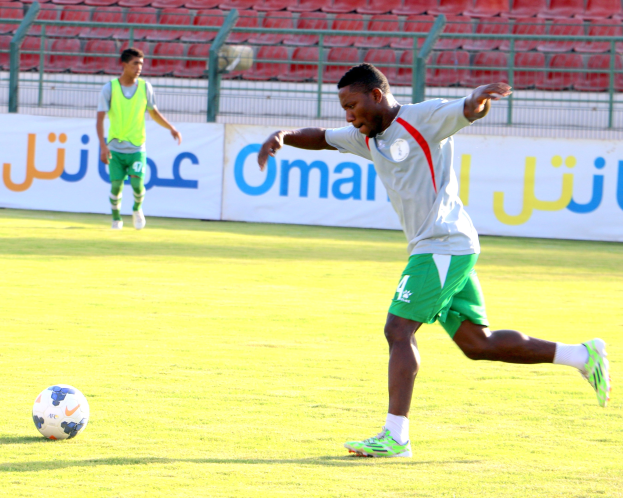 Ein Mann, der einen Fußball auf einem Rasenfeld in einem Stadion mit zwei Personen in der Nähe und Bannern, Geländern und Stühlen im Hintergrund kickt.