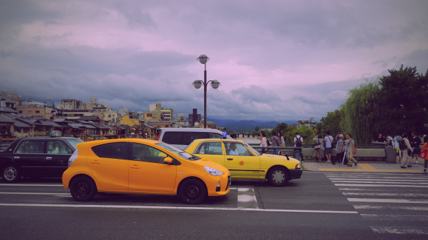 Zwei gelbe Taxis stehen am Straßenrand mit Fußgängern auf dem Bürgersteig, Laternenpfählen, Bäumen, Gebäuden und einem bewölkten Himmel im Hintergrund.