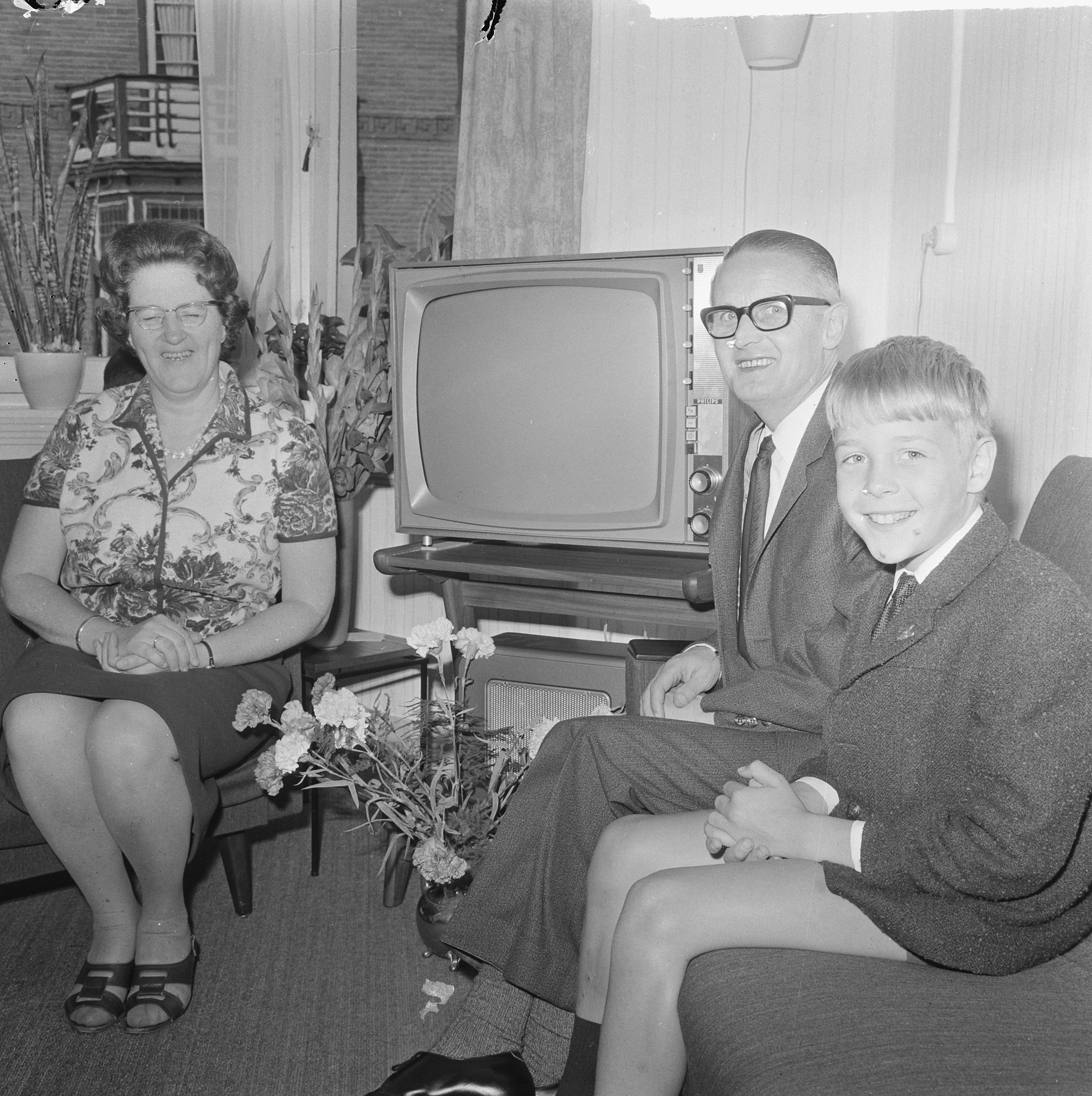 Ein Schwarz-Weiß-Foto eines Mannes, einer Frau und eines Jungen, die auf einem Sofa vor einem Fernseher sitzen, mit Blumenvasen auf einem Tisch daneben und Vorhängen im Hintergrund.
