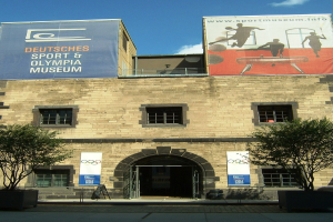 Außenansicht des Deutschen Sport & Olympischen Museums in Berlin, das ein Gebäude mit Fenstern und einer Tür zeigt, das von Bäumen und Bannern umgeben ist, unter einem bewölkten Himmel.
