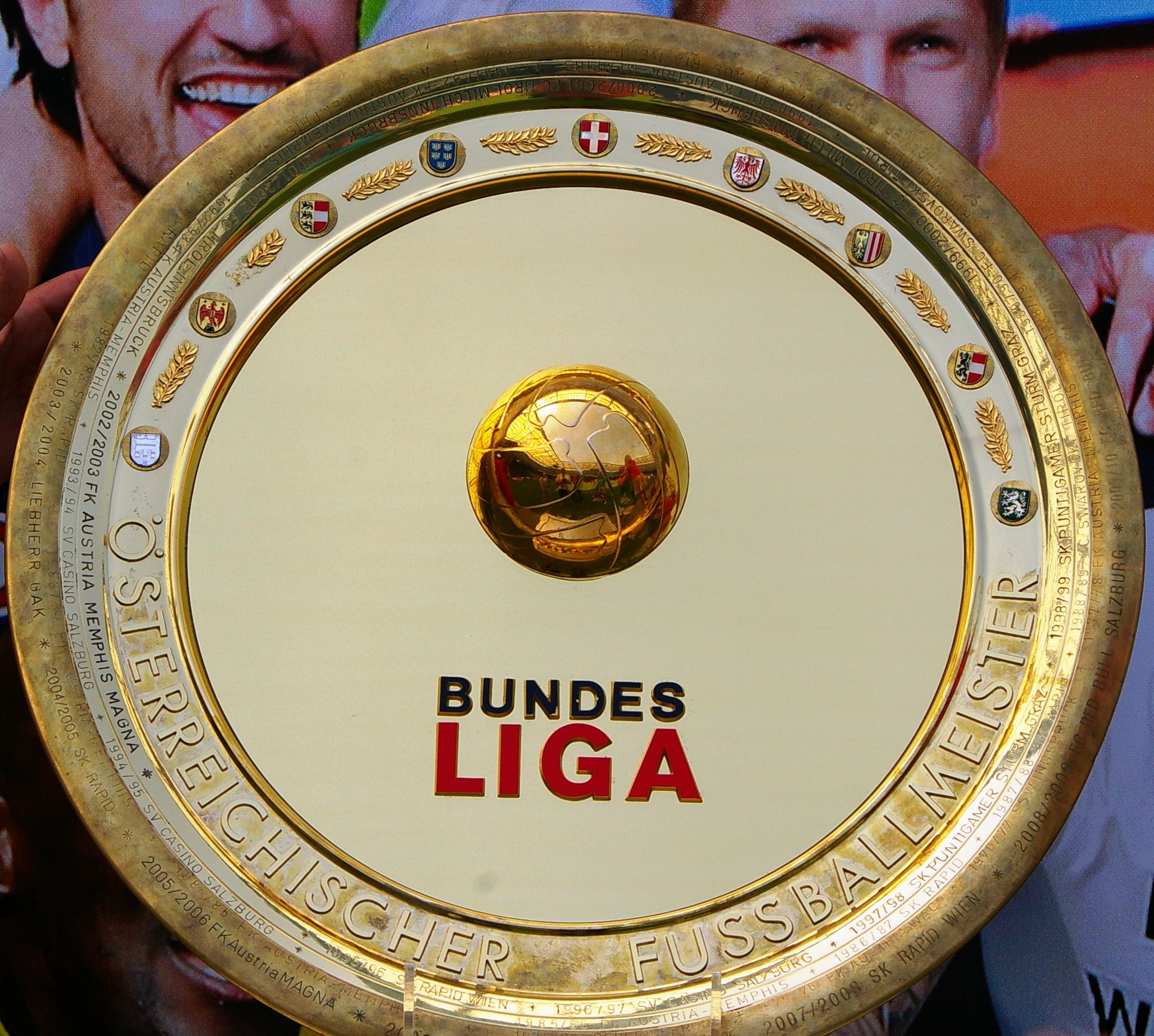 Goldener Schild mit 'Bundesliga'-Text, gehalten von einem Mann mit einem Banner im Hintergrund mit drei Personen.