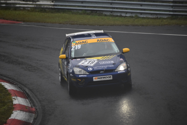 Ein blaues Rennauto mit Text und Zahlen darauf fährt eine Strecke entlang, flankiert von Gras und Geländern, mit Bäumen im Hintergrund.