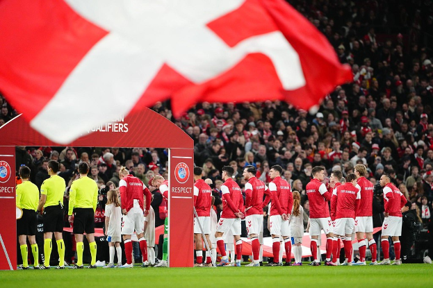 Eine Gruppe von Menschen auf einem Fussballfeld mit einer roten und weissen Flagge im Vordergrund, einem Bogen mit Text "Bayern München vs Bayern München Betting Tips & Preview" im Hintergrund und einer grossen Menge im Stadion.