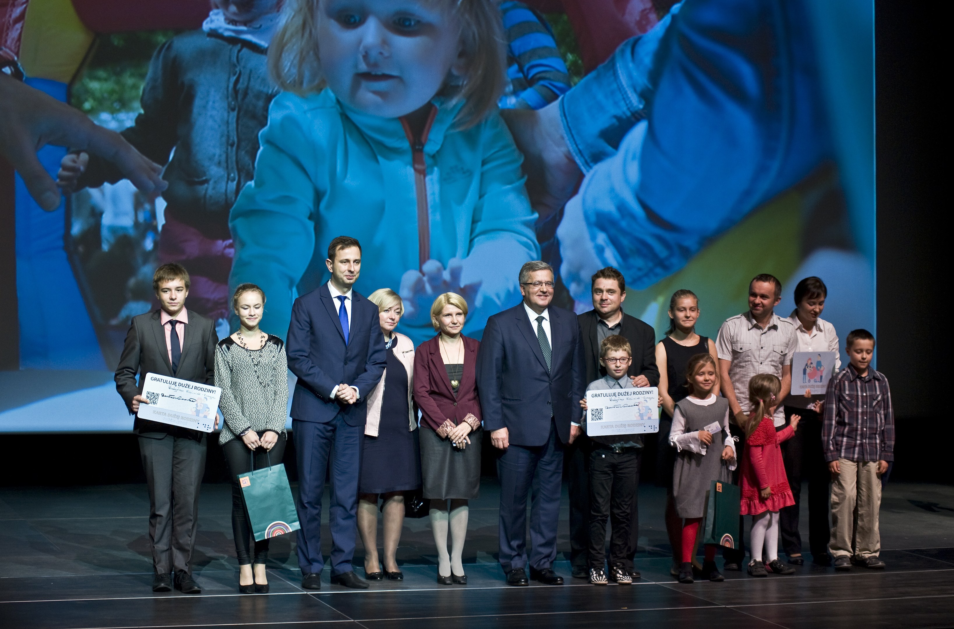 Eine Gruppe von Menschen auf einer Bühne mit Taschen und Urkunden, im Hintergrund ein Bildschirm mit einem Bild eines Kindes und anderen Personen.