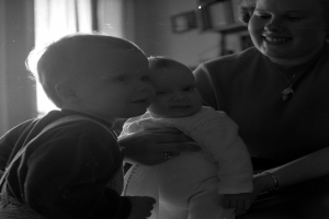 Eine Frau hält ein Baby mit einem warmen Lächeln in den Armen, während zwei andere Kinder neben ihr stehen, in einem gemütlichen Raum mit Rahmen und einem Fenster im Hintergrund.