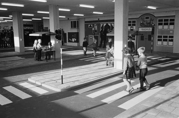 Schwarzes und weißes Foto von Kindern, die eine Straße an einer Ampel überqueren, mit Fußgängern in der Nähe, einem Verkehrssignalmast im Vordergrund und architektonischen Elementen im Hintergrund.