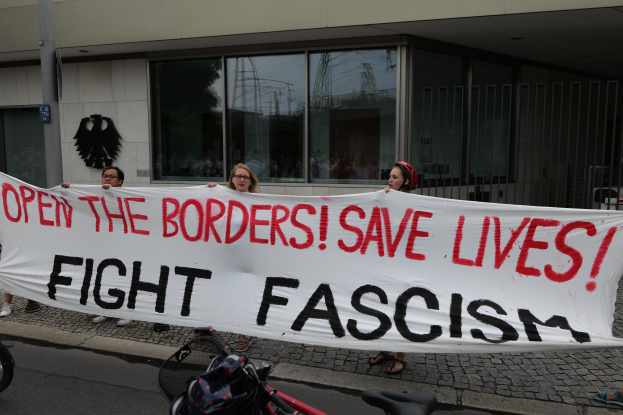 Eine Gruppe von Menschen hält ein Transparent mit der Aufschrift "Öffnet die Grenzen, rettet Leben, kämpft gegen Faschismus" vor einem Gebäude mit Glasfenstern und einem Mast im Hintergrund, mit einem Fahrrad und einer Tasche auf der Straße.